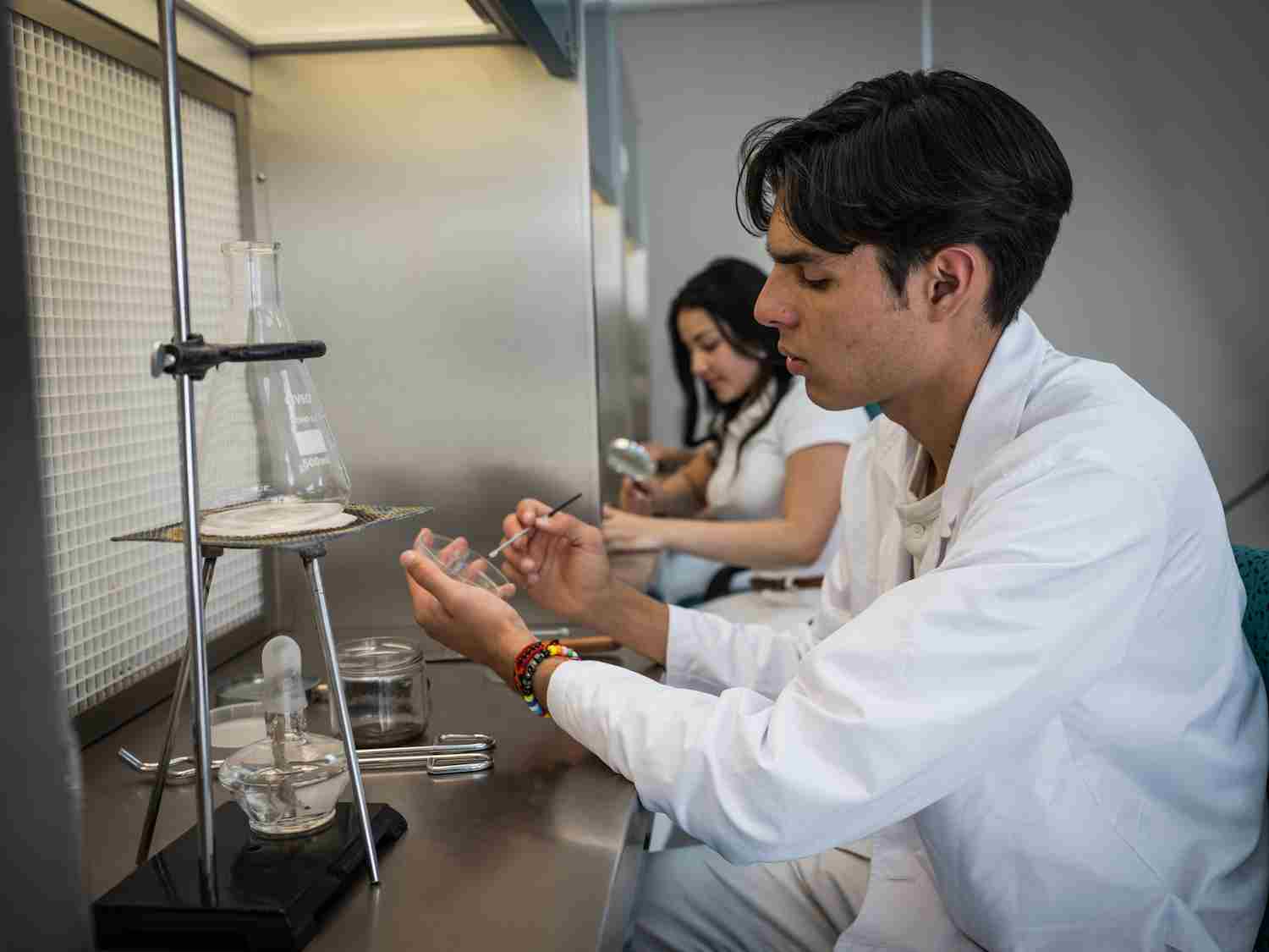 Estudiante en bata blanca manipula material de laboratorio junto a un mechero y un matraz en campana de extracción.